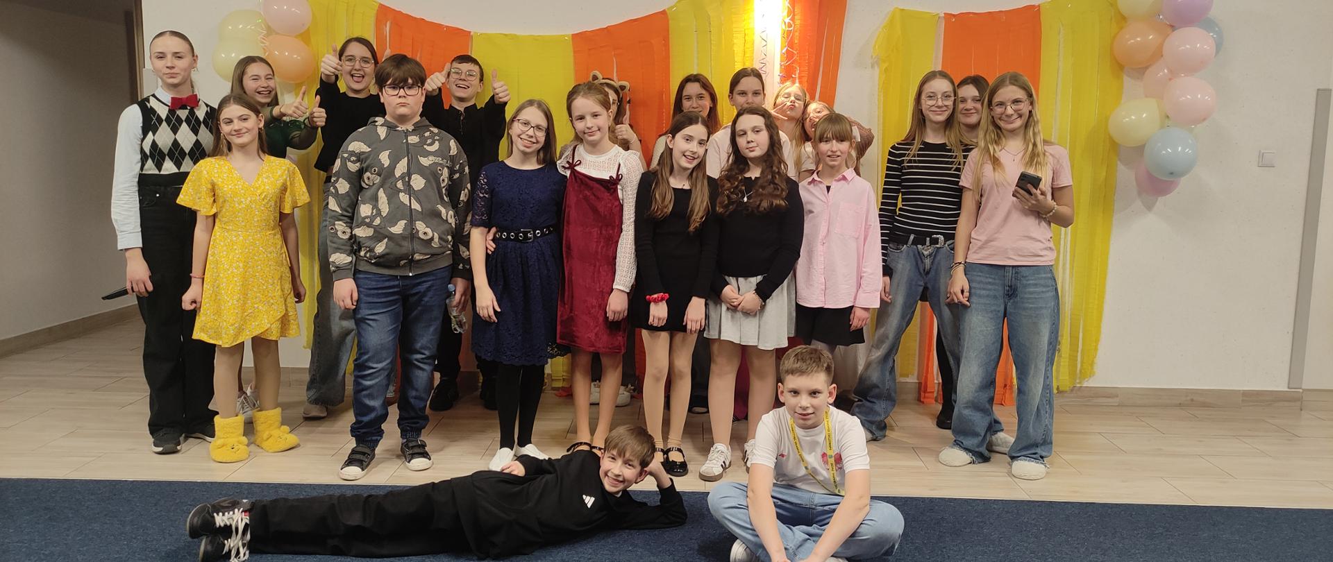 Uczniowie pozują do wspólnego zdjęcia na tle karnawałowych dekoracji, uśmiechają się i pokazują gesty radości, tworząc pogodną i przyjazną atmosferę wydarzenia