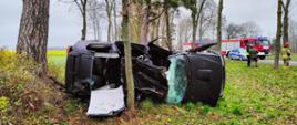 Uszkodzone auto leży na boku na poboczu drogi. W aucie wycięto część dachu. W tle samochody ratowniczo-gaśnicze i radiowóz.
