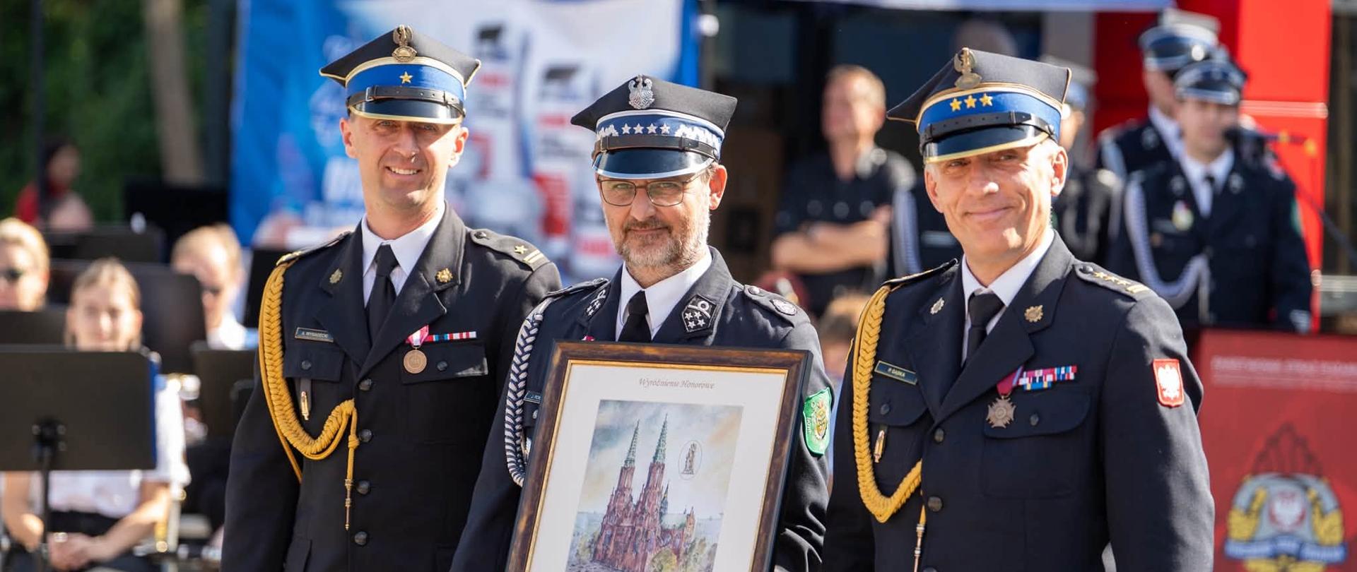 W centrum kadru stoją trzej strażacy i zaproszeni goście w galowych mundurach OSP z rogatywkami na głowach oraz odznaczeniami na piersiach. Mężczyzna w środku trzyma w rękach oprawiony w elegancką, ciemną ramę obraz przedstawiający zabytkowy kościół lub strażnicę, przekazany jako pamiątkowy dar jubileuszowy. W tle widać niebiesko-białą konstrukcję namiotu eventowego oraz zieleń otaczających drzew. Pogoda jest pełna słońca i światła dziennego. Zdjęcie promienieje radością, wdzięcznością, serdecznością oraz dumą z pielęgnowania strażackiej historii.