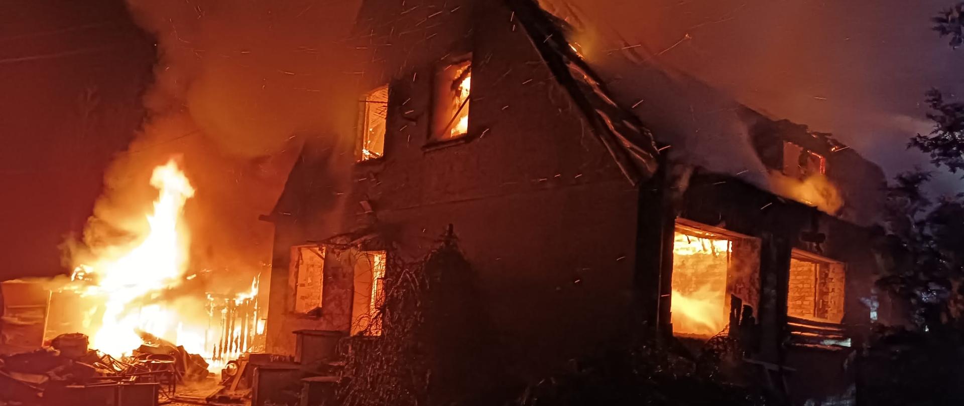 Płonący budynek mieszkalny. Ogień trawi całe jego wnętrze wraz z dachem. W górę unoszą się kłęby dymu. Ogniem objęta jest również stojąca przy obiekcie wiata.