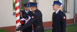 Plac przed komendą. Poczet flagowy w umundurowaniu wyjściowym stoi przed biało-czerwonym masztem. Flagowy wraz z dowódcą pocztu przymocowują flagę do masztu. Obok nich w pozycji zasadniczej stoi strażak asytujący podnosi flagę. W tle zieleń i budynek komendy.