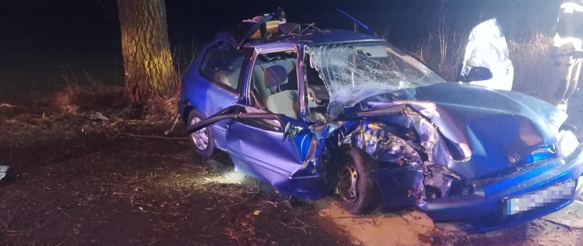 Zdjęcie przedstawia rozbity samochód osobowy koloru niebieskiego, po uderzeniu w drzewo. Rozbity prawy przód pojazdu, uszkodzenia drzwi i głównej szyby. Plamy wyciekłych płynów pod pojazdem zasypane sorbentem.