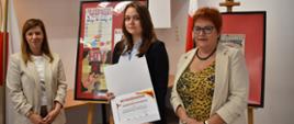 Trzy osoby stoją obok siebie. Osoba znajdująca się w centrum trzyma otwartą teczkę z dyplomem z widocznym nagłówkiem „Wyróżnienie” oraz torbę z upominkami. Po lewej i prawej stronie stoją dwie osoby w jasnych marynarkach. W tle znajdują się sztalugi z oprawionymi plakatami edukacyjnymi dotyczącymi HIV/AIDS i profilaktyki zdrowotnej.