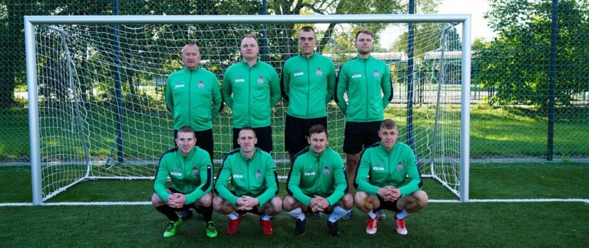 Zdjęcie przedstawia drużynę w zielonych dresach sportowych. Siedmiu zawodników ustawiło się przed bramką – czterech stoi, a trzech kuca w pierwszym rzędzie. Każdy z piłkarzy ma czarną lub czerwoną odzież sportową na nogach i obuwie piłkarskie. W tle widoczne są drzewa i ogrodzenie boiska. Zawodnicy pozują spokojnie, patrząc wprost w obiektyw.