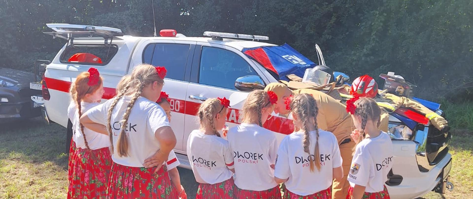 Dziewczęta w strojach ludowych stoją przed maską wozu strażackiego i przyglądają się strażakowi, który pokazuje im rozłożony sprzęt gaśniczy i czerwony hełm.
