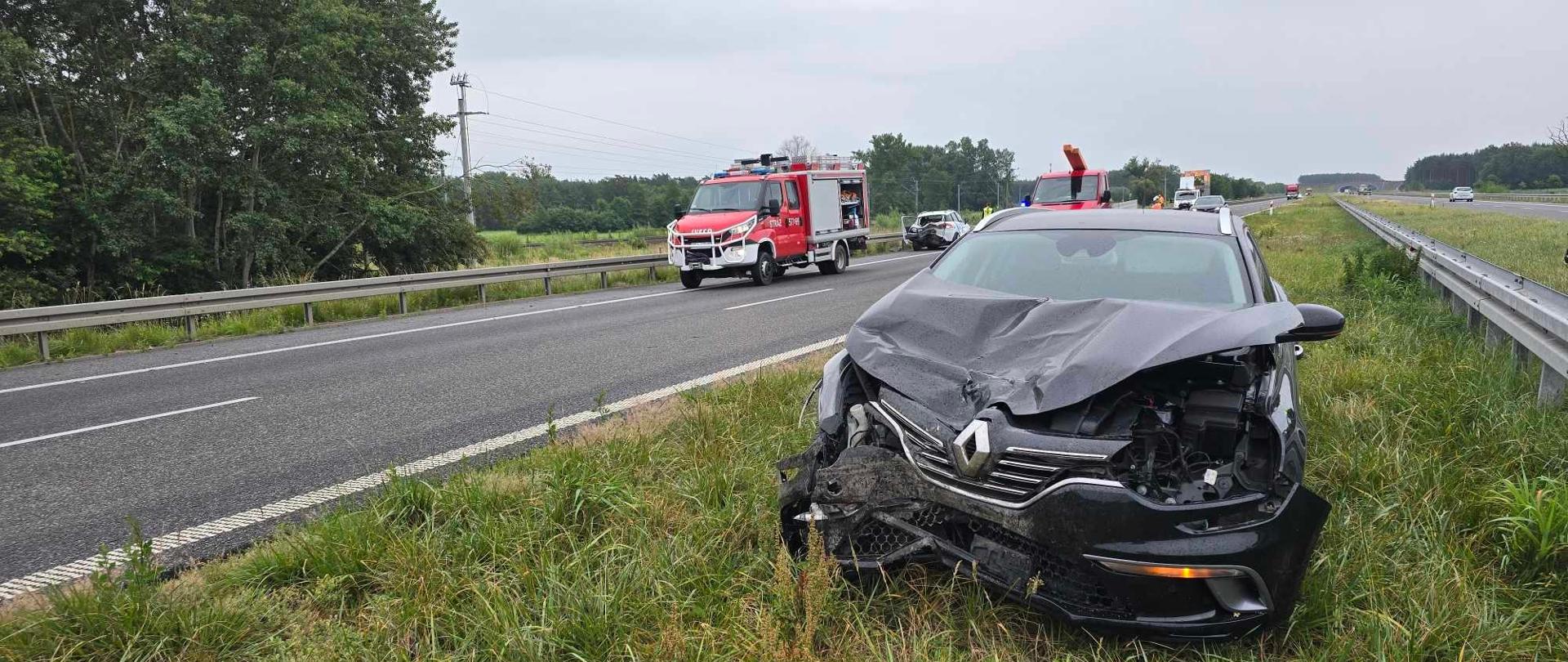 Na pasie zieleni stoi czarne auto z rozbitym przodem. Po lewej stronie na jezdi - wóz strażacki oraz drugie auto uczestniczące w kolizji