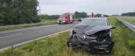 Na pasie zieleni stoi czarne auto z rozbitym przodem. Po lewej stronie na jezdi - wóz strażacki oraz drugie auto uczestniczące w kolizji