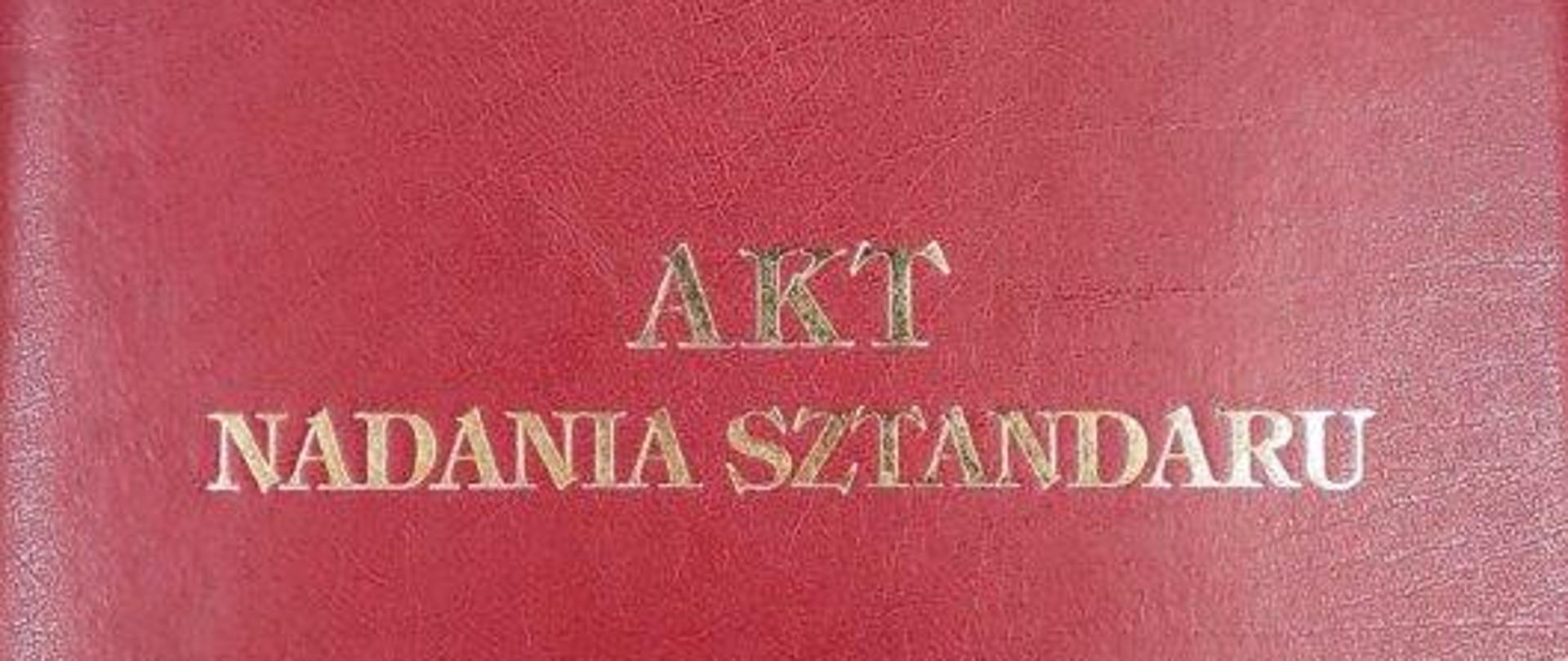 Na zdjęciu widzimy ozdobną okładkę do aktu nadania sztandaru. Okładka koloru czerwonego, na środku okładki na czerwonym tle naniesione jest godło państwowe, złoty orzeł w koronie. Poniżej złoty napis :"AKT NADANIA SZTANDARU". 