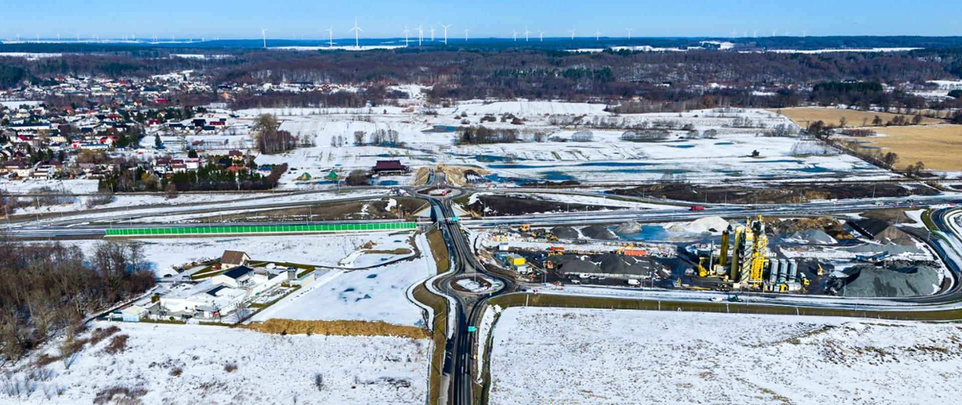Zdjęcie lotnicze zimowego krajobrazu przedstawiające węzeł drogowy w budowie. Widać kilka skrzyżowań i rond, obszerny plac budowy z maszynami i instalacjami oraz pokryte śniegiem pola i tereny leśne. Po lewej stronie znajduje się zabudowa miejscowości, a w tle widoczne są wiatraki elektrowni wiatrowej.