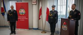 Zdjęcie zrobione w pomieszczeniu strażnicy na tle na tle godła Rzeczpospolitej Polskiej, Sztandaru Komendy Powiatowej Państwowej Straży Pożarnej w Lublińcu, dwóch Flag Rzeczpospolitej Polskiej oraz baneru Komendy Powiatowej PSP w Lublińcu. Na pierwszym planie, po lewej stronie funkcjonariusz odchodzący na zaopatrzenie emerytalne, po prawej stronie Komendant Powiatowy Państwowej Straży Pożarnej w Lublińcu st. bryg. Janusz Bula stojący za mównicą oraz pomiędzy nim a flagami Rzeczpospolitej Polskiej dowódca uroczystości. Strażacy ubrani w umundurowanie galowe.