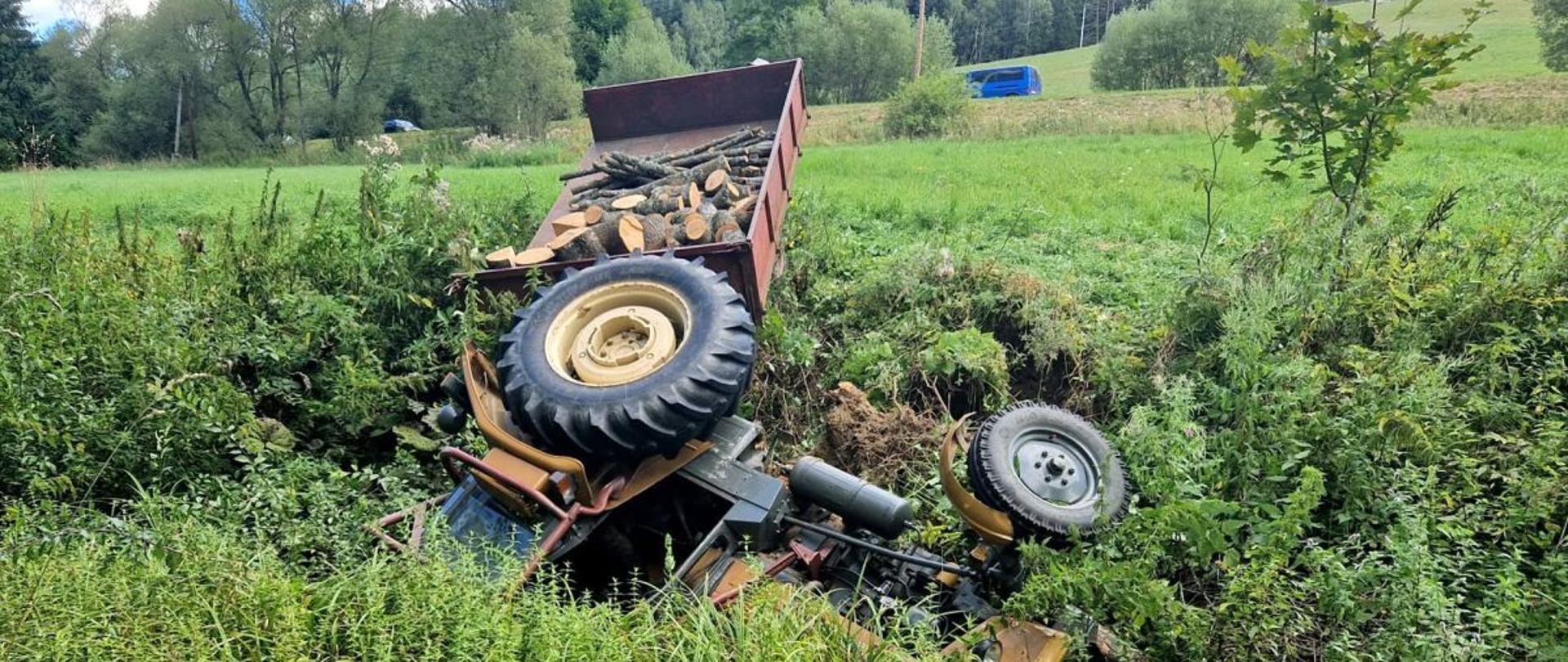 Zdjęcie przedstawia ciągnik rolniczy leżący na boku po wypadku.