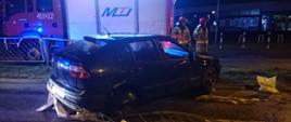 Uszkodzony samochód osobowy który uderzył w barierki przydrożne. W tle samochód gaśniczy i dwóch strażaków zabezpieczających teren zdarzenia.