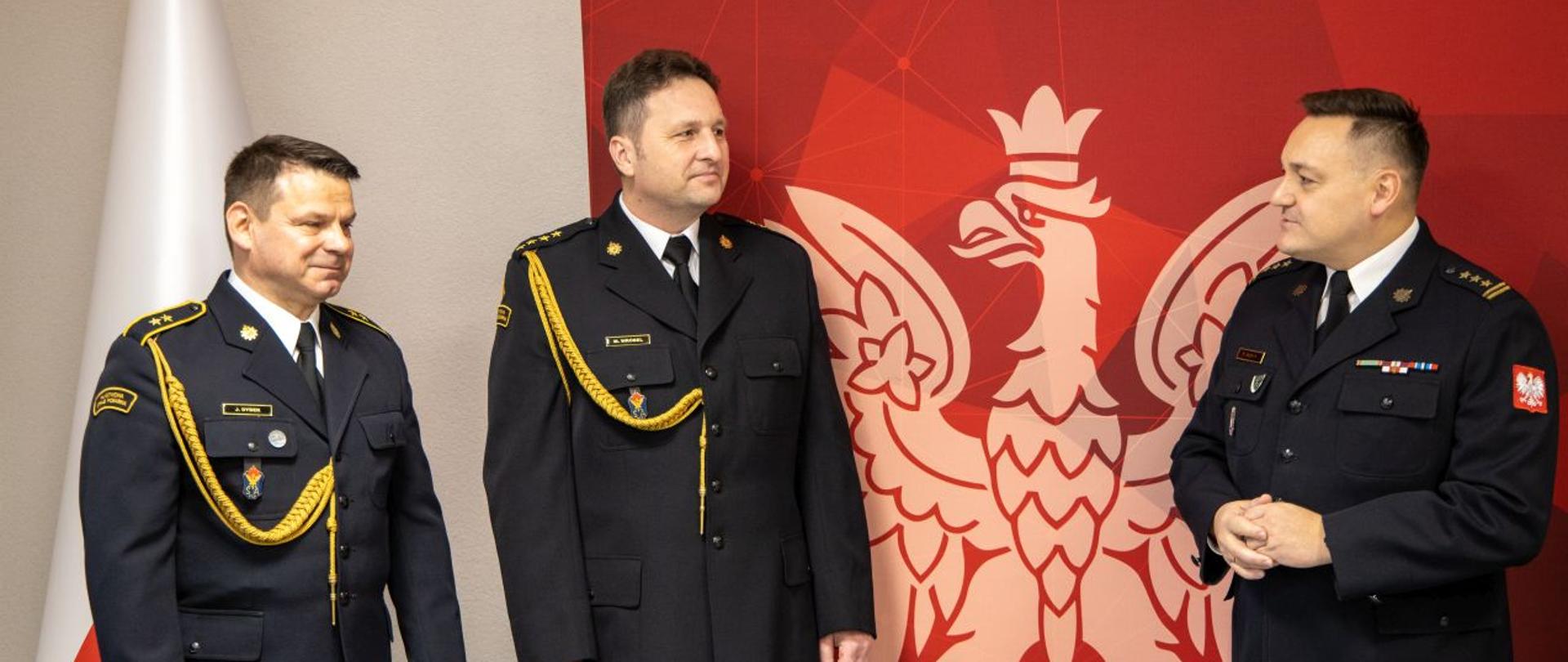 Dwóch strażaków KW PSP w Opolu przechodzących na zaopatrzenie emerytalne: st. kpt. Mirosław Wróbel i st. asp. Janusz Dybek w umundurowaniu galowym wysłuchują przemówienia Opolskiego Komendanta Wojewódzkiego PSP w Opolu st. bryg. Krzysztofa Kędryka. W tle widoczny czerwony banner Państwowej Straży Pożarnej Komendy Wojewódzkiej PSP w Opolu z jasnym Godłem RP oraz biało czerwoną Flagą RP.