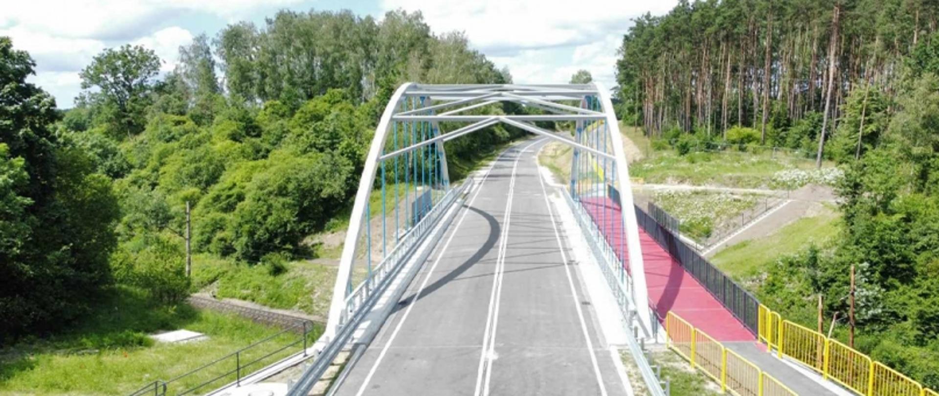 Most z metalową konstrukcją łukową nad drogą, z czerwonym chodnikiem dla pieszych lub rowerzystów i żółtymi barierkami, otoczony zielenią.