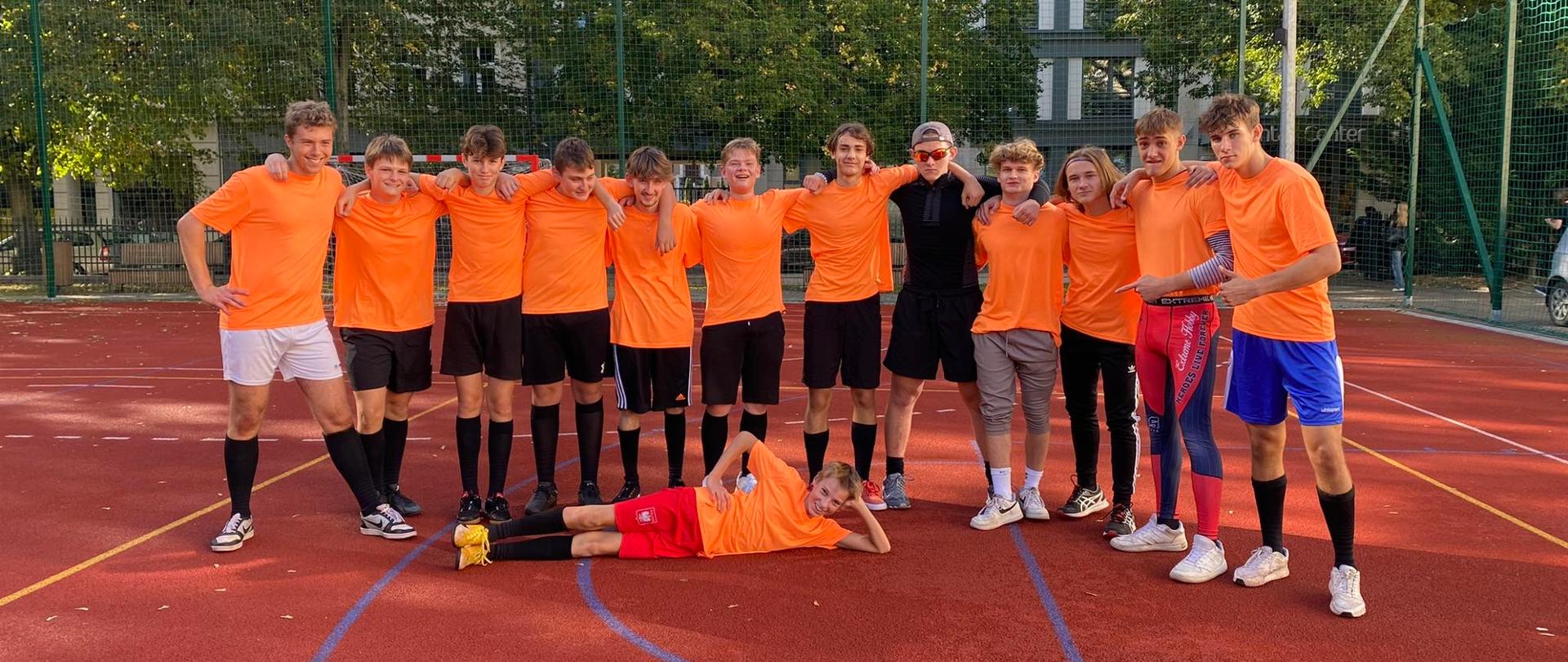 zdjęcie przedstawia drużynę w pomarańczowych koszulkach podczas wspólnego pozowanego zdjęcia po meczu, jeden z zawodników leży na boisku