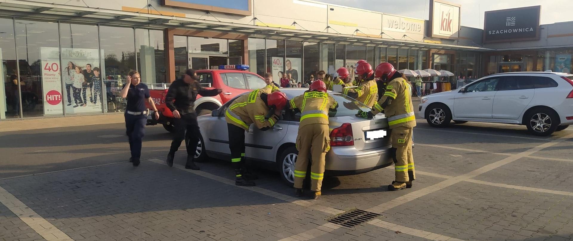 Dziecko zatrzaśnięte w aucie. Parking przed jedną z galerii handlowych w Rawiczu. Na parkingu zaparkowane są dwa samochody osobowe. Wokół jednego z nich znajduje się 7 strażaków oraz policjantka. Strażacy próbują otworzyć zatrzaśnięte drzwi. sześciu z nich ma na sobie ubrania specjalne koloru piaskowego i czerwone hełmy. W tle terenowe auto strażackie oraz budynek galerii.