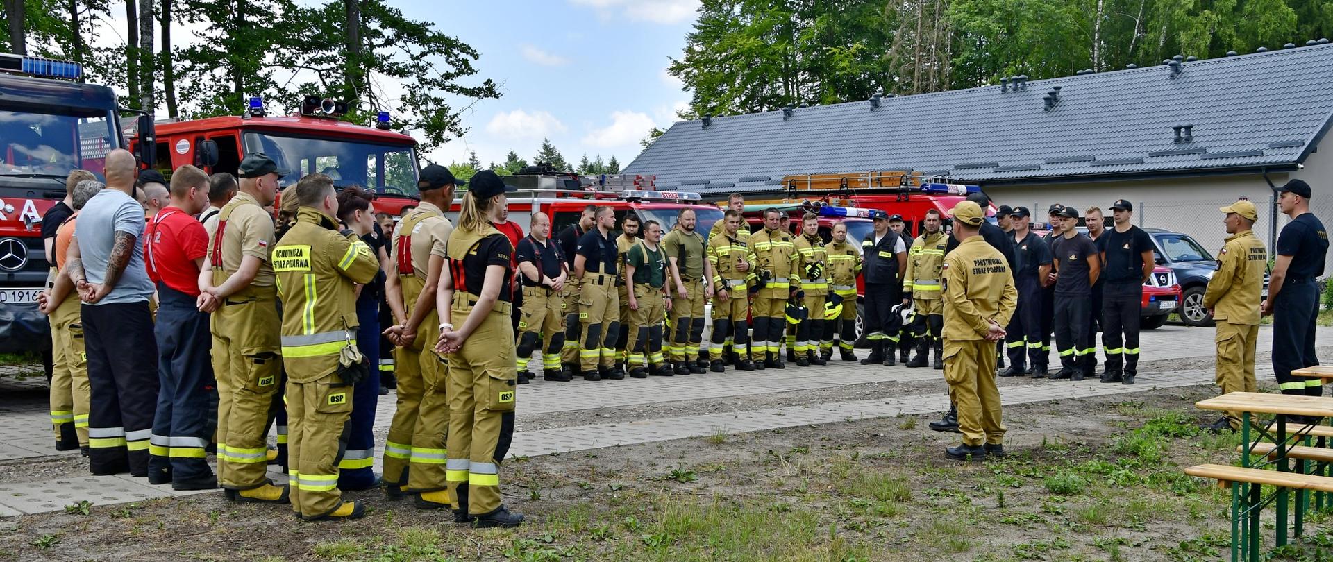 Zdjęcie przedstawia zbiórkę po ćwiczeniach na lasach. Strażacy stoją ustawieni w litere U i słuchaja podsumowania.