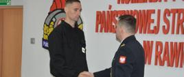 Świetlica komendy powiatowej. Na tle ściany, na której widnieje czerwony napis Komenda Powiatowa Państwowej Straży Pożarnej w Rawiczu i logo PSP, komendant powiatowy ściskając dłoń składa gratulacje jednemu z nowych strażaków. 