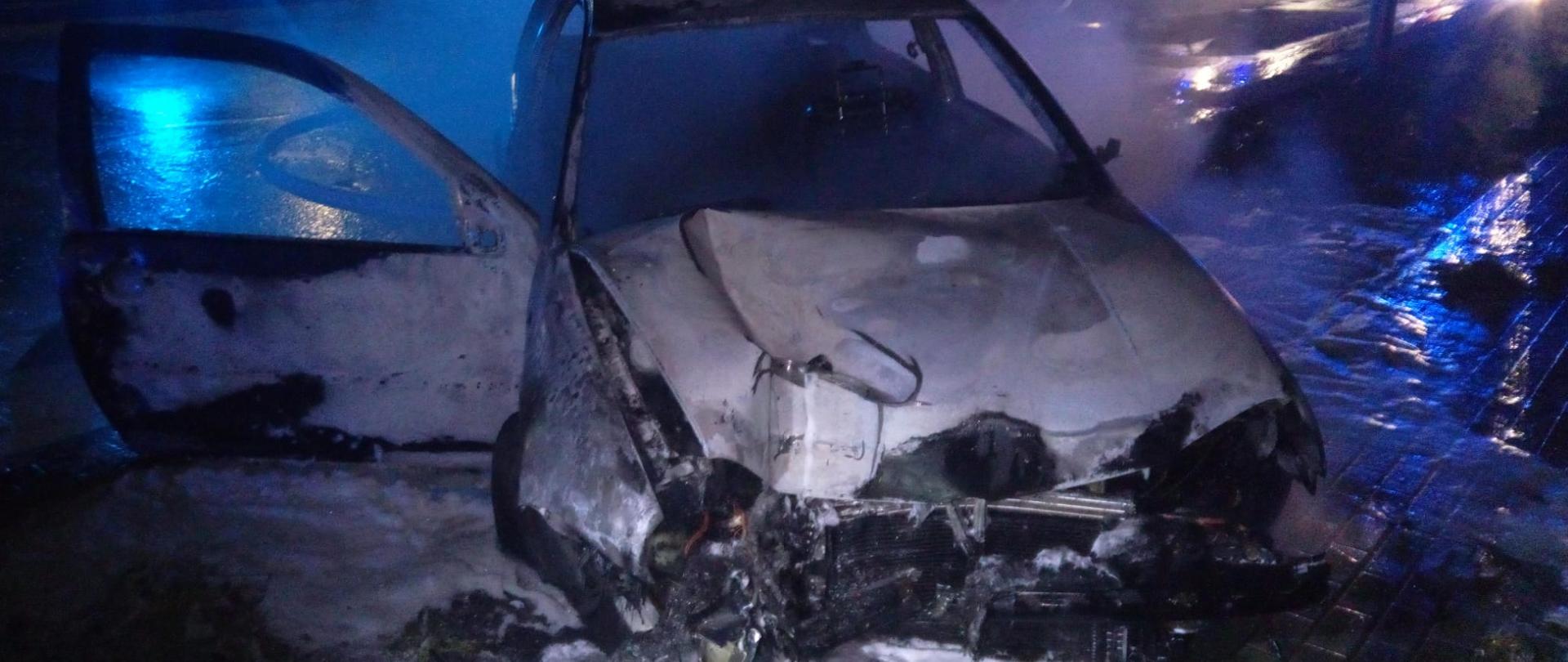 Zdjęcie przedstawia spalony samochód osobowy. Przedni pas zniszczony w wyniku uderzenia w słup latarni ulicznej. Wokół rozlana piana gaśnicza.