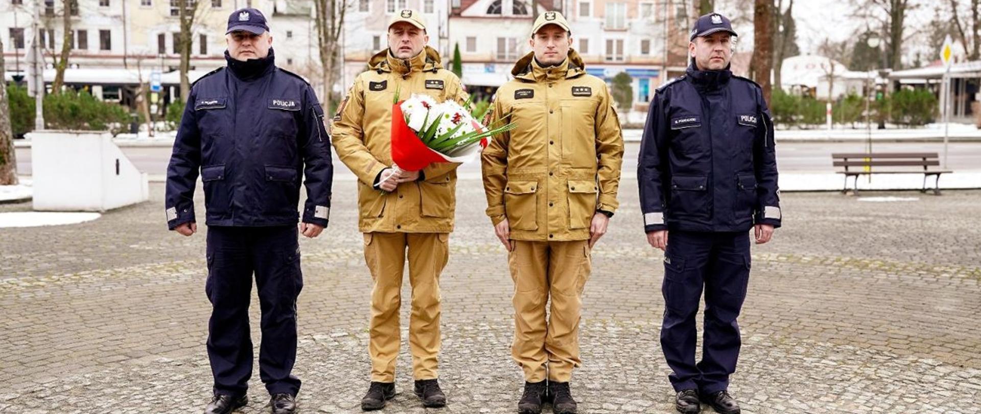 Zdjęcie przedstawia Komendantów Powiatowych Policji w Ustrzykach Dolnych oraz Komendanta Powiatowego Państwowej Straży Pożarnej w Ustrzykach Dolnych i kwatermistrza ustrzyckiej straży pożarnej. Wszyscy wymienieni stoją na rynku w Ustrzykach Dolnych. Komendant straży pożarnej trzyma kwiaty. 