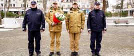 Zdjęcie przedstawia Komendantów Powiatowych Policji w Ustrzykach Dolnych oraz Komendanta Powiatowego Państwowej Straży Pożarnej w Ustrzykach Dolnych i kwatermistrza ustrzyckiej straży pożarnej. Wszyscy wymienieni stoją na rynku w Ustrzykach Dolnych. Komendant straży pożarnej trzyma kwiaty. 