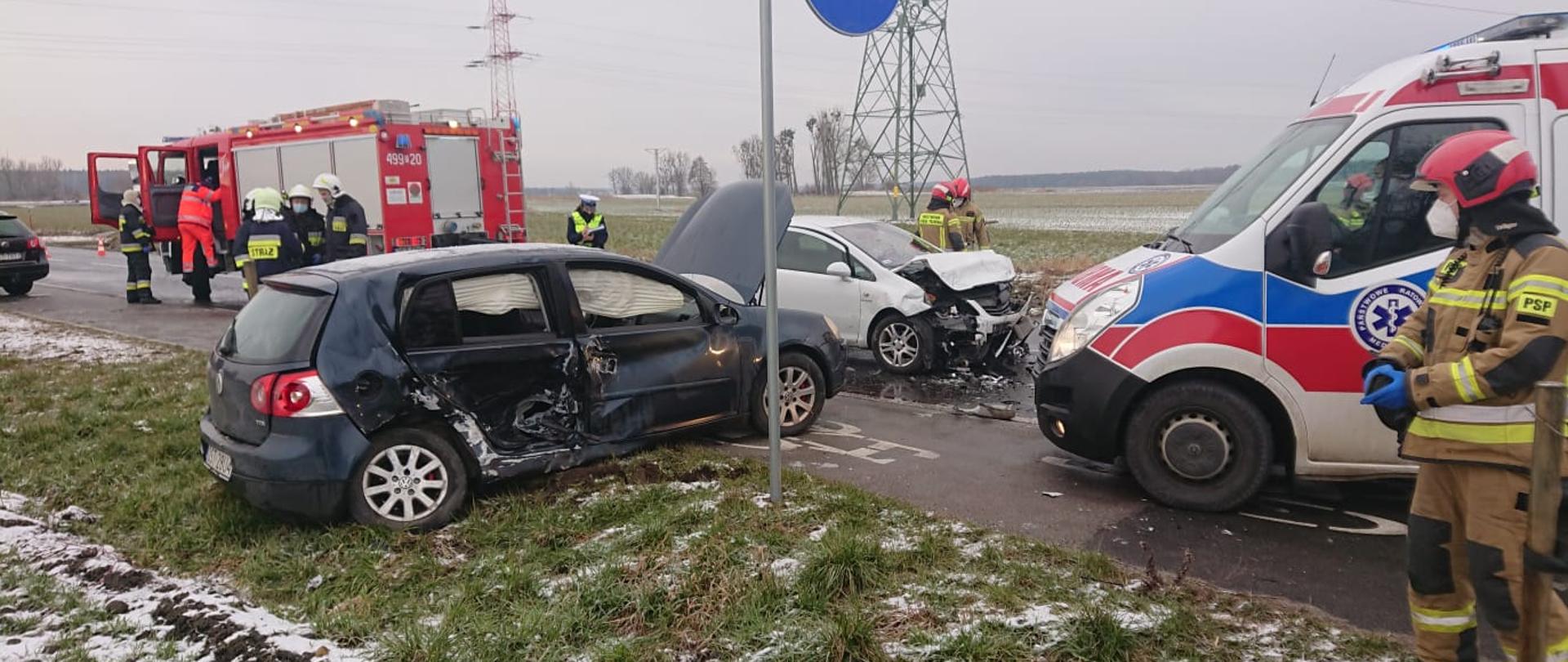 Zdjęcie przedstawia dwa rozbite samochody znajdujące się na jezdni, karetkę pogotowia, samochód pożarniczy i strażaków.