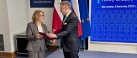 Przewodnicząca Rady Służby Publicznej Małgorzata Janyska odbiera akt powołania z rąk min. Jakuba Stefaniaka, sekretarza stanu, Zastępcy Szefa Kancelarii Prezesa Rady Ministrów; w tle flagi Polski i Unii Europejskiej