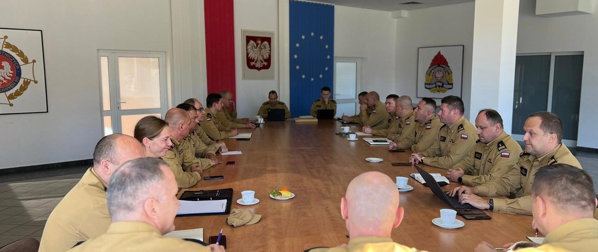 Zdjęcie przedstawia odprawę strażaków w jasnej sali konferencyjnej. Przy długim stole siedzi około 16-20 osób. Wszyscy uczestnicy ubrani są w jednolite piaskowe mundury. Sala jest jasna i przestronna, ze świetlówkami umieszczonymi w kasetonowym suficie. Ściany są białe. Na centralnej ścianie, za głową stołu, wiszą dwie duże flagi oraz godło. Od lewej: Flaga w kolorach polskich (czerwień i biel). Pomiędzy flagami, na białym tle, wisi Godło Rzeczypospolitej Polskiej (biały orzeł w koronie). Flaga Unii Europejskiej (niebieskie tło z dwunastoma żółtymi gwiazdami).