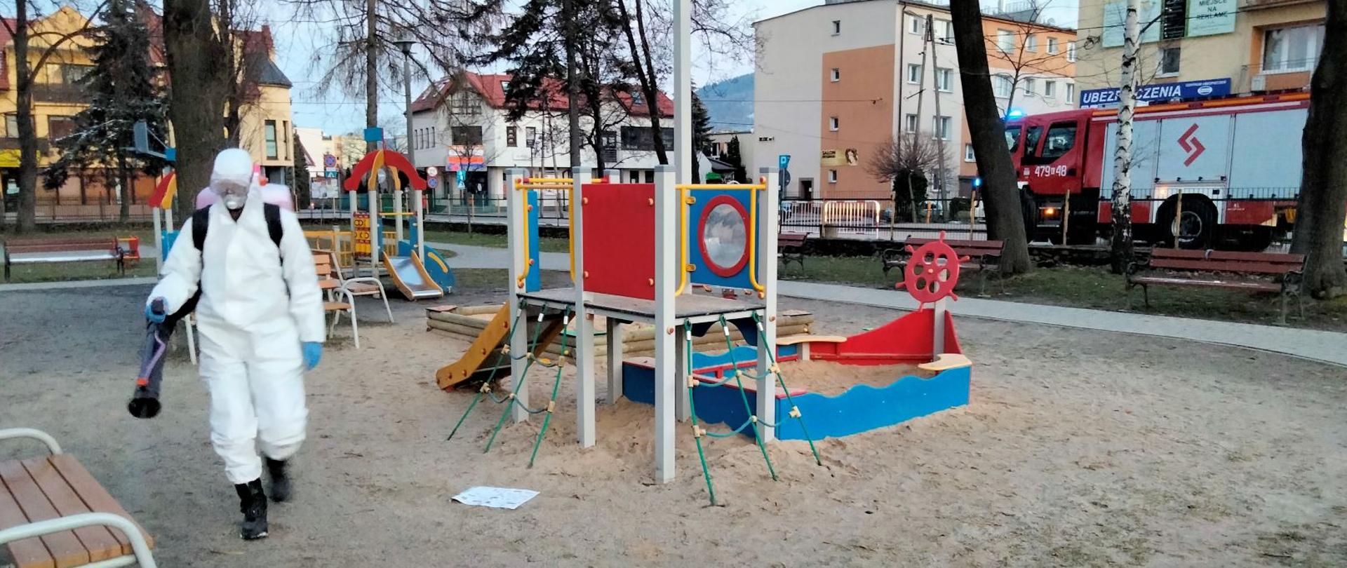 W środkowej części zdjęcia widoczne elementy infrastruktury placu zabaw. Z lewej strony strażak zabezpieczony w kombinezon, maskę i gogle przeprowadza dezynfekcję wykorzystując opryskiwacz spalinowy. Po prawej stronie na zdjęciu widoczny za ogrodzeniem samochód ratowniczo - gaśniczy.