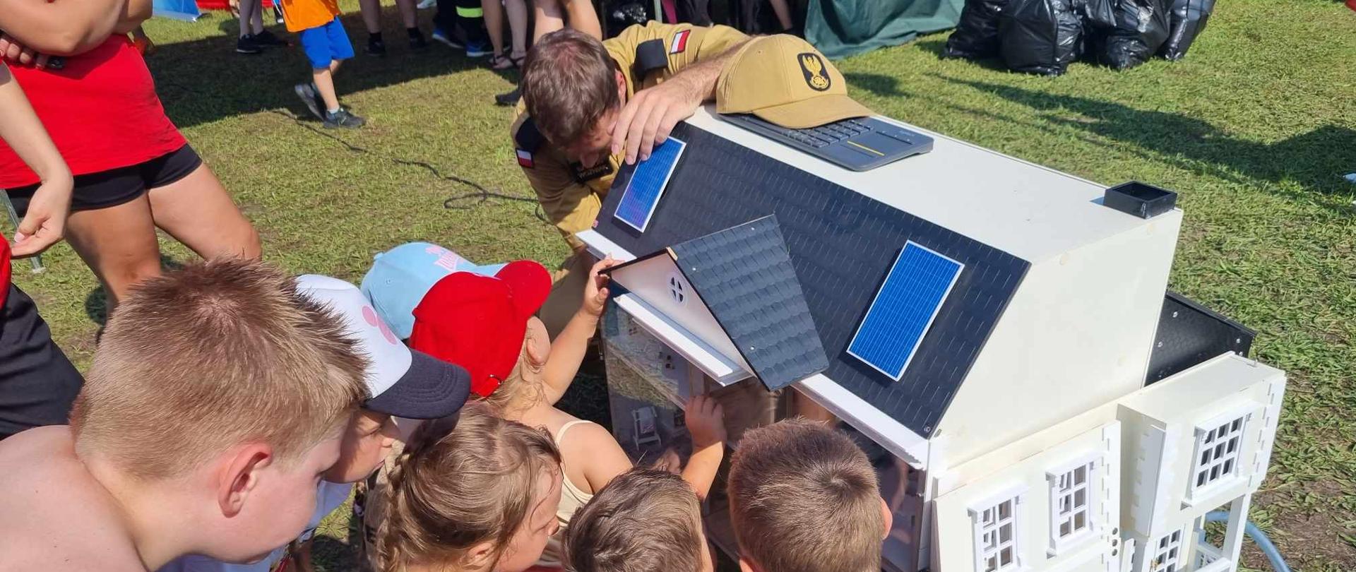Biwak na Leśnym. Fotografia przedstawia fragment boiska. Na pierwszym planie gromadka dzieci stojąca przy symulatorze zagrożeń pożarowych (dom jednorodzinny koloru białego). Obok symulatora znajduje się strażak, który opowiada i pokazuje dzieciom zagrożenia występujące w domu.