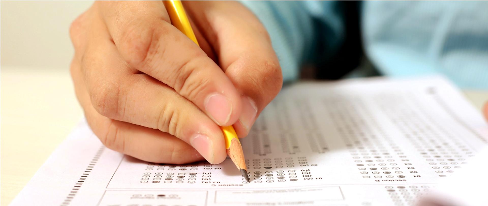 Grafika przedstawia dłoń osoby wypełniającej arkusz egzaminacyjny