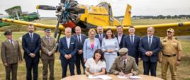 Podpisanie umowy na dofinansowanie lotów patrolowo - gaśniczych. Podpisania dokonuje Pani Aleksandra Drescher Prezes WFOŚiGW w Opolu i Dyrektor RDLP w Katowicach. W tle zaproszeni goście a za nimi samolot gaśniczy Dromader.