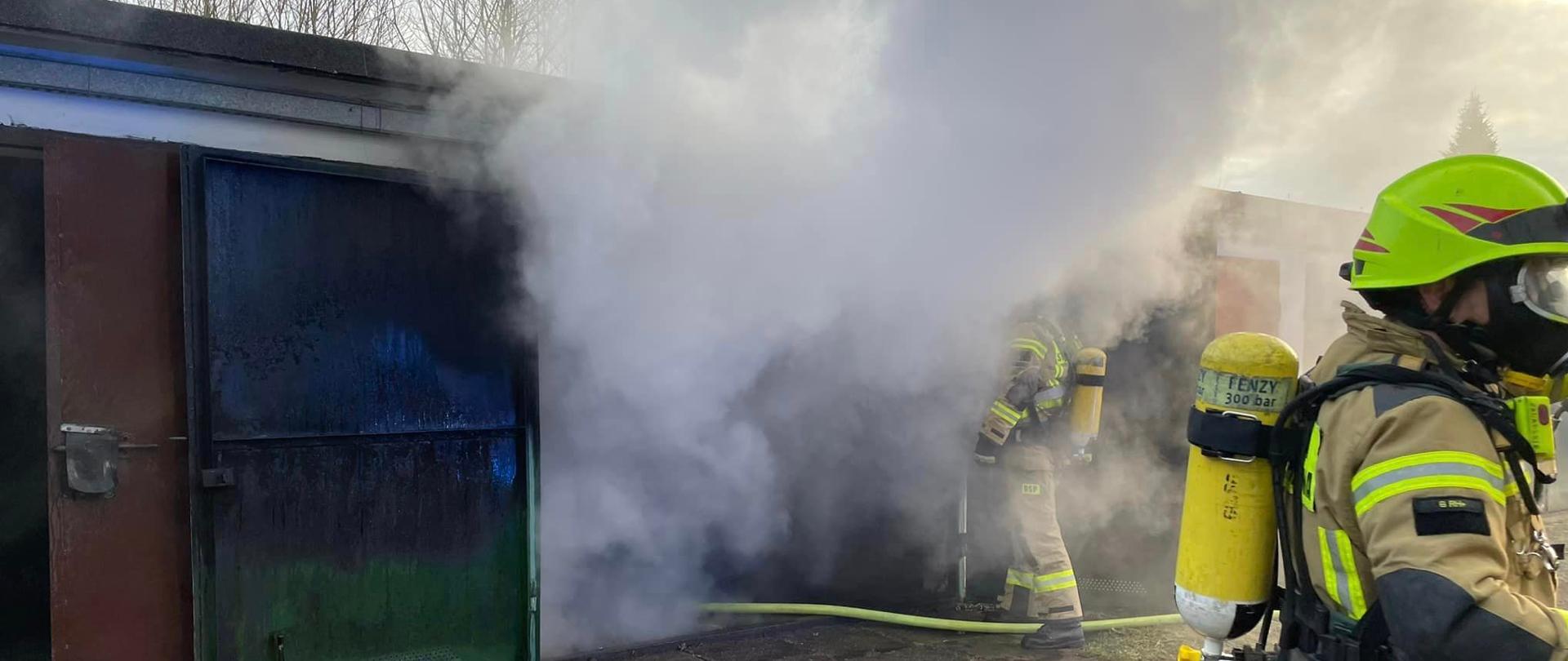 Na zdjęciu znajdują się strażacy w ubraniach specjalnych na tle garaży