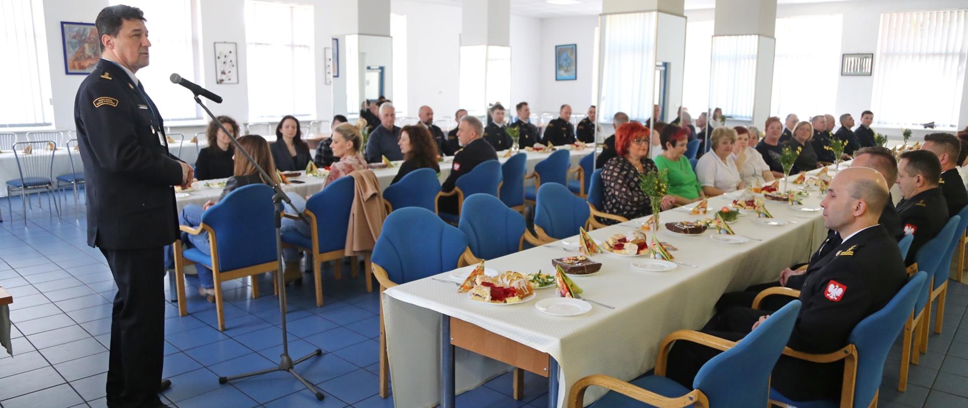 Komendant Centralnej Szkoły PSP, w umundurowaniu galowym, przemawia stoją przed mikrofonem. W głębi widzimy pracowników Szkoły siedzących przy stolach, podczas spotkania wielkanocnego