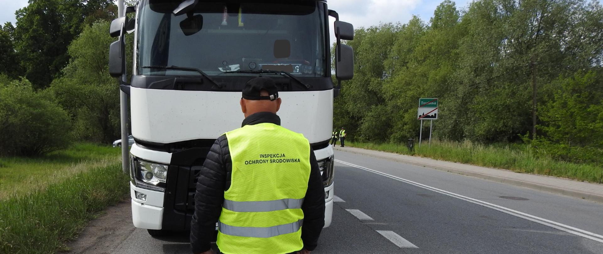 Mężczyzna obrócony plecami do zdjęcia, ubrany w długie spodnie, kurtkę, czapkę oraz odblaskową kamizelkę z napisem Inspekcja Ochrony Środowiska. Przed mężczyzną stoi samochód ciężarowy. Zdjęcie zostało zrobione na drodze, dookoła są drzewa i trawy. 