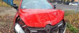 Widać, czerwony, uszkodzony pojazd. Zniszczenia w przedniej części pojazdu. Zachmurzenie duże. W tle inne pojazdy oraz rośliny. 