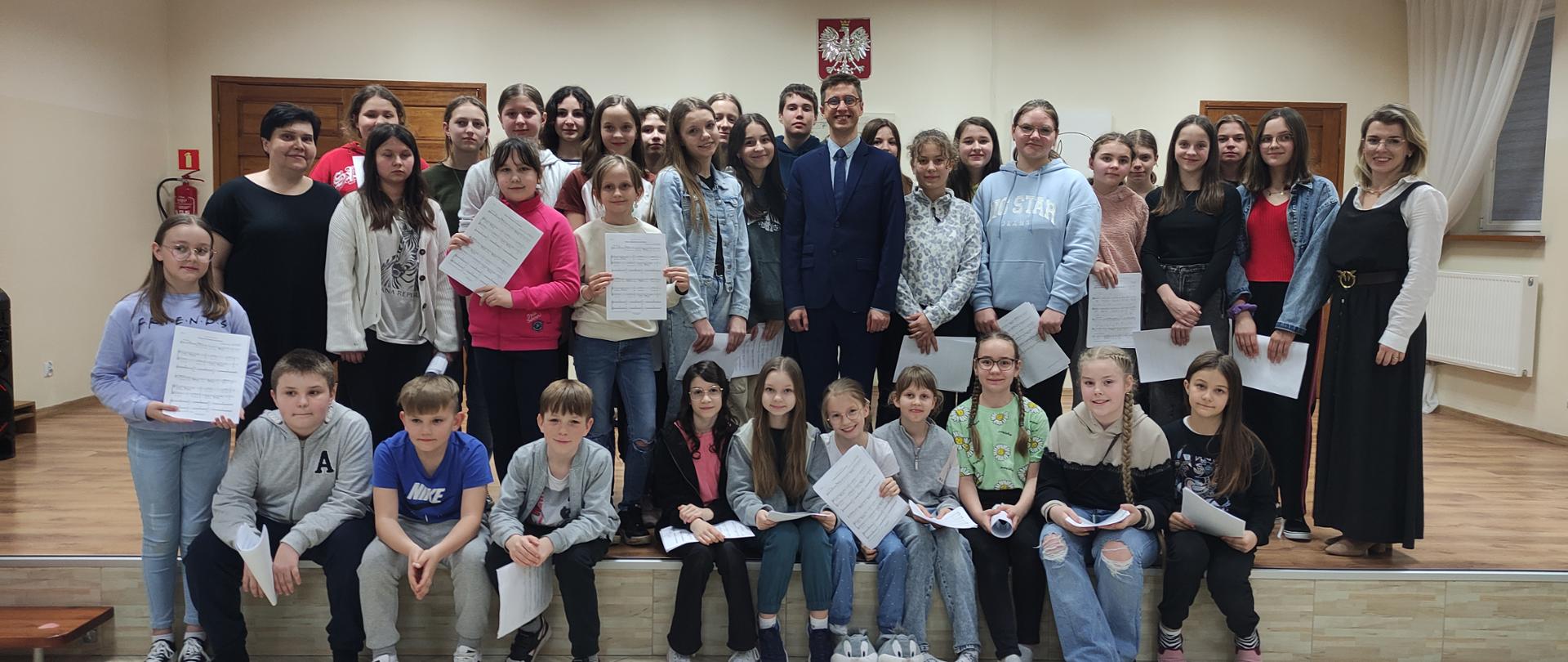 Grupa około 30 osób stoi na scenie. Część z nich trzyma w dłoniach kartki z nutami. W tle na ścianie godło. 