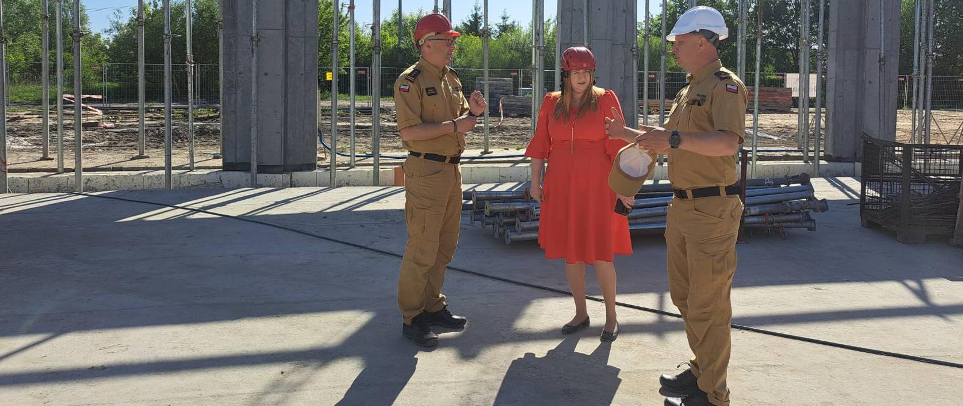 Kobieta w czerwonej sukni i dwóch mężczyzn w piaskowych mundurach stojący i rozmawiający wewnątrz budowanego obiektu.