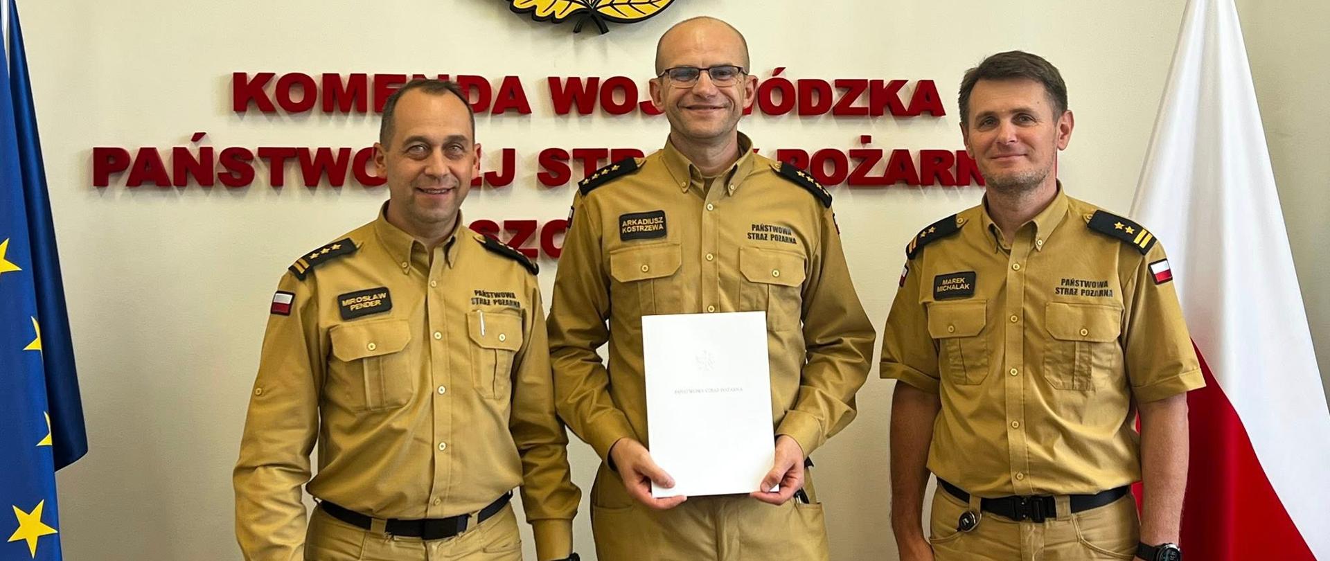 Na zdjęciu widać trzech mężczyzn w mundurach straży pożarnej w kolorze piaskowym. Stoją na tle ściany, na której widnieje duży emblemat Państwowej Straży Pożarnej z orłem w koronie, płomieniem i liśćmi dębu oraz napis „KOMENDA WOJEWÓDZKA PAŃSTWOWEJ STRAŻY POŻARNEJ”. Po lewej stronie widoczna jest flaga Unii Europejskiej, a po prawej – flaga Polski na srebrnym maszcie zakończonym grotami. Mężczyzna w środku trzyma w dłoniach biały dokument. Wszyscy trzej uśmiechają się i stoją obok siebie.