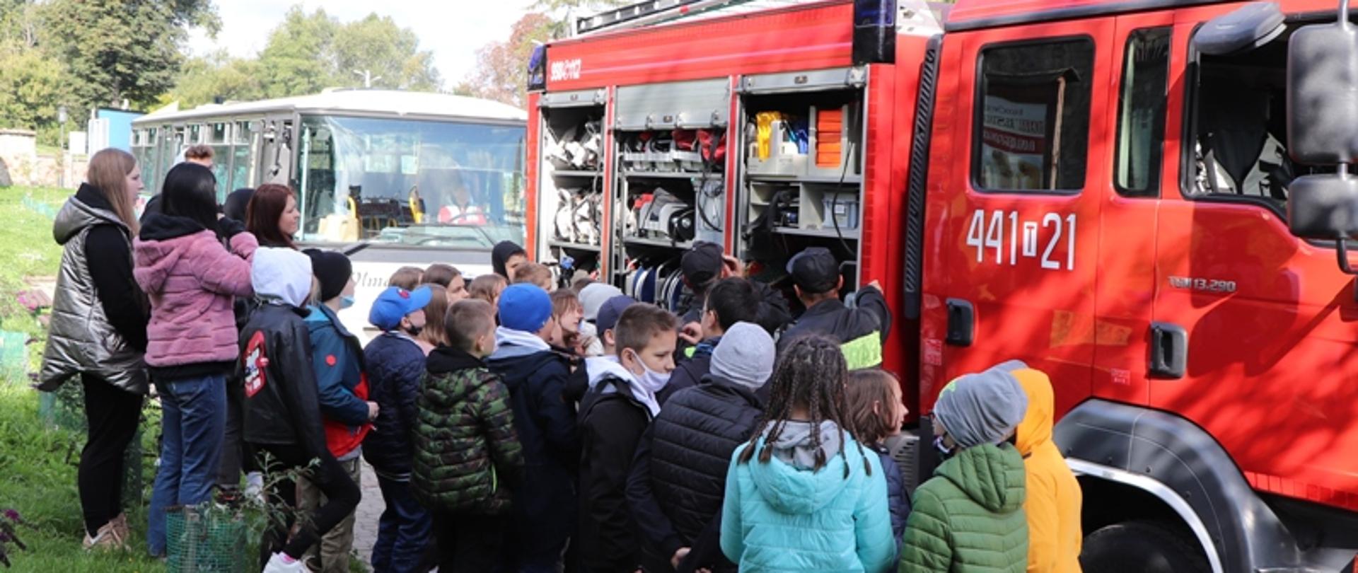 Zdjęcie przedstawia czerwony samochód pożarniczy oraz grupę dzieci podczas pogadanki przy sprzęcie pożarniczym