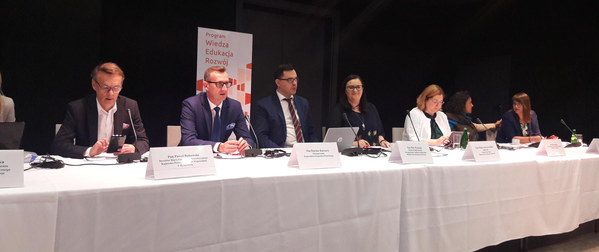 Na zdjęciu długi stół przykryty białym materiałem, przy stole siedzi siedem osób, za nimi roll-up z napisem "Program Wiedza Edukacja Rozwój"