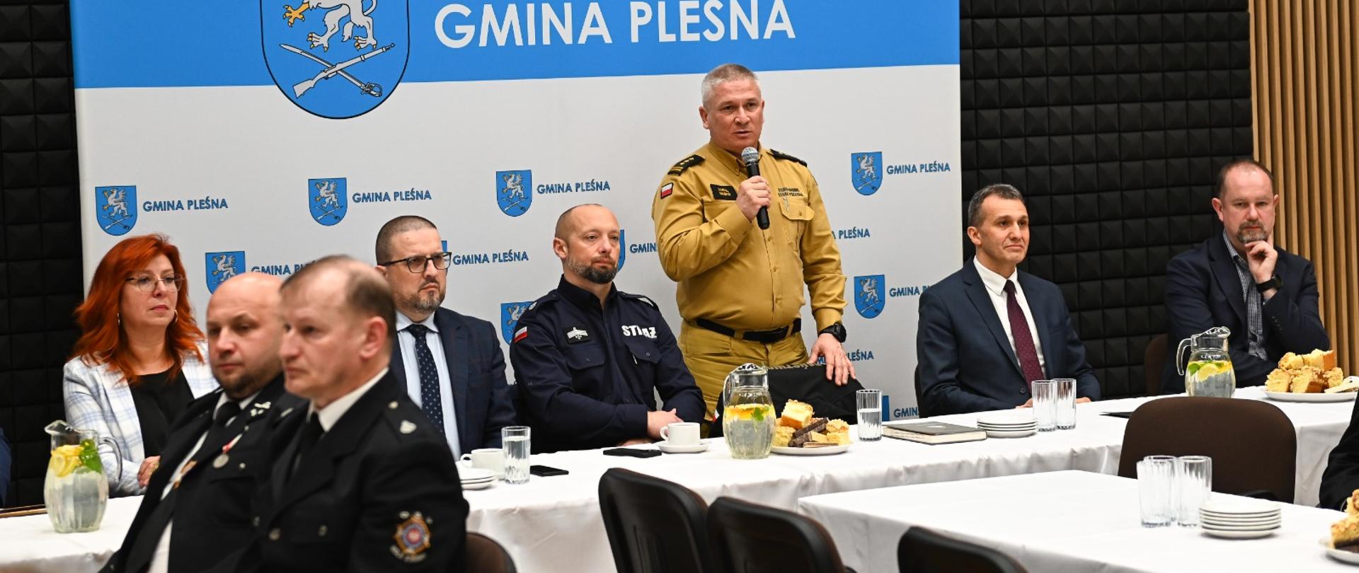 Przed banerem Gmina Pleśna przy stołach siedzą strażacy i zaproszeni goście . Na środku za stołem stoi komendant ubrany w piaskowy mundur w ręku trzyma mikrofon. Stoły nakryte są białymi obrusami.