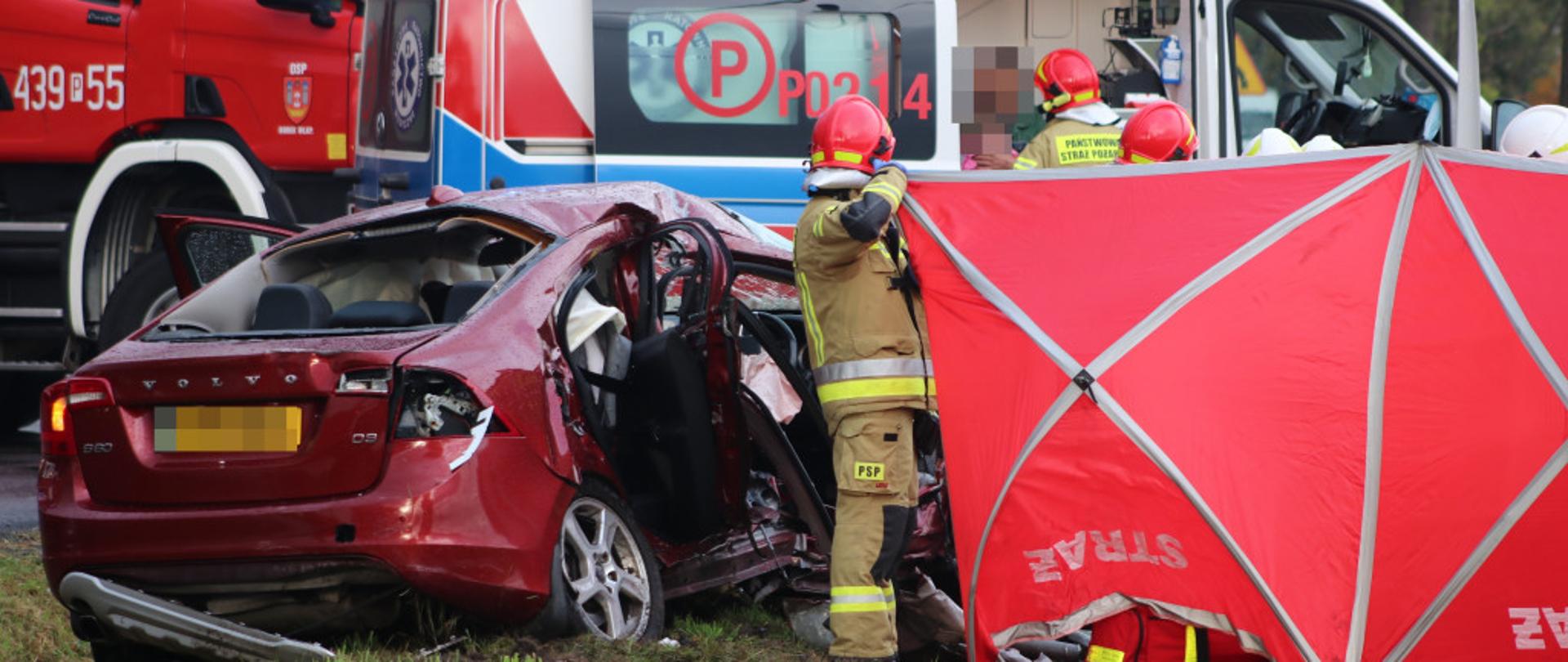Zdjęcie przedstawia uszkodzony samochód osobowy oraz strażaków trzymających parawan. W tle widać karetkę pogotowia oraz samochód strażacki. 