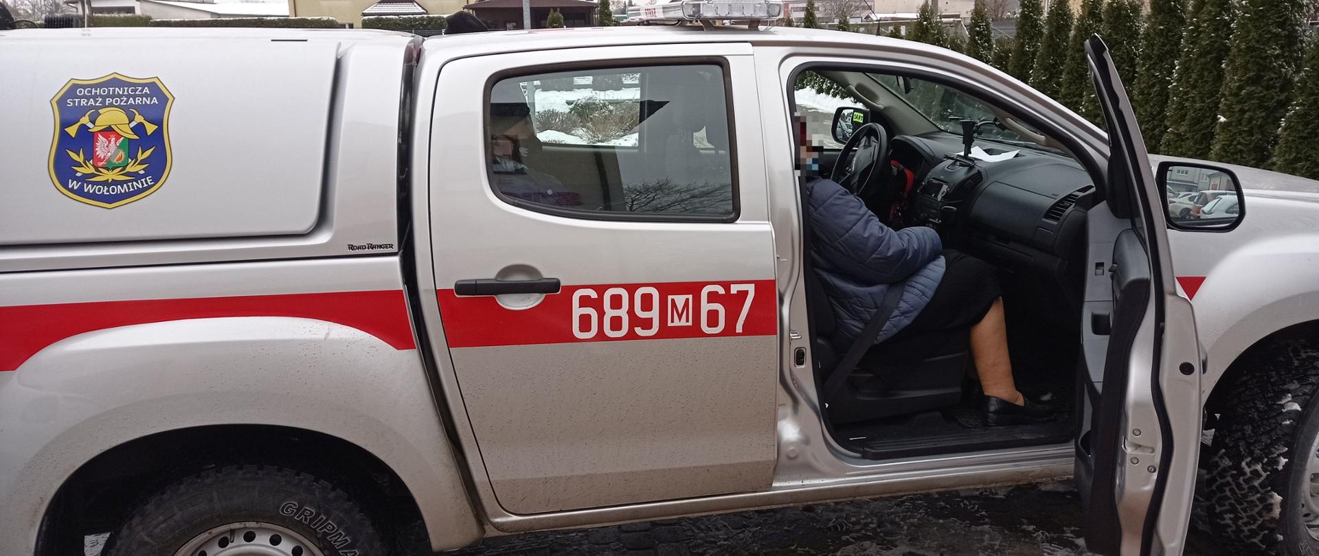 Na zdjęciu widoczny srebrny samochód strażacki z czerwonym pasem. W środku widać kobietę przewiezioną do punktu szczepień. Drzwi pojazdu są otwarte i kobieta przygotowuje się aby opuścić pojazd.