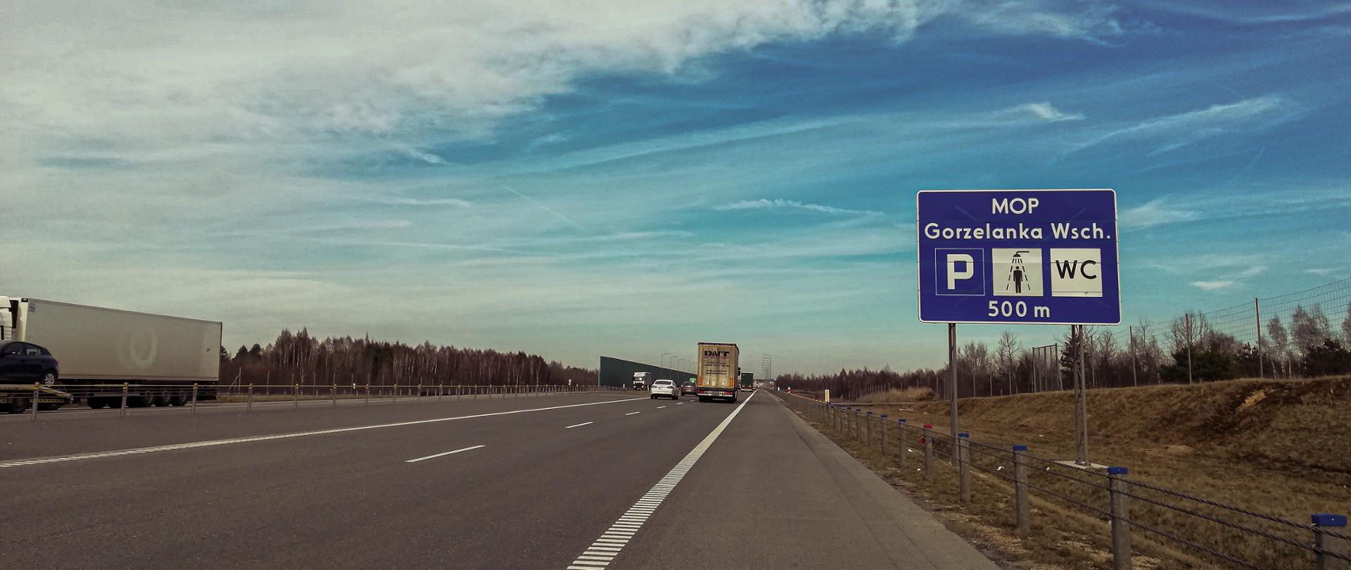Fotografia z poziomu terenu autostrady A1. W prawej części fotografii znajduje się tablica informacyjna, złożona z trzech znaków drogowych na niebieskim oraz informacją MOP Gorzelanka Wchód. Znaki informują o parkingu, prysznicu, WC. W lewej i środkowej części fotografii zlokalizowana jest dwujezdniowa autostrada. Po autostradzie poruszają się pojazdy. 