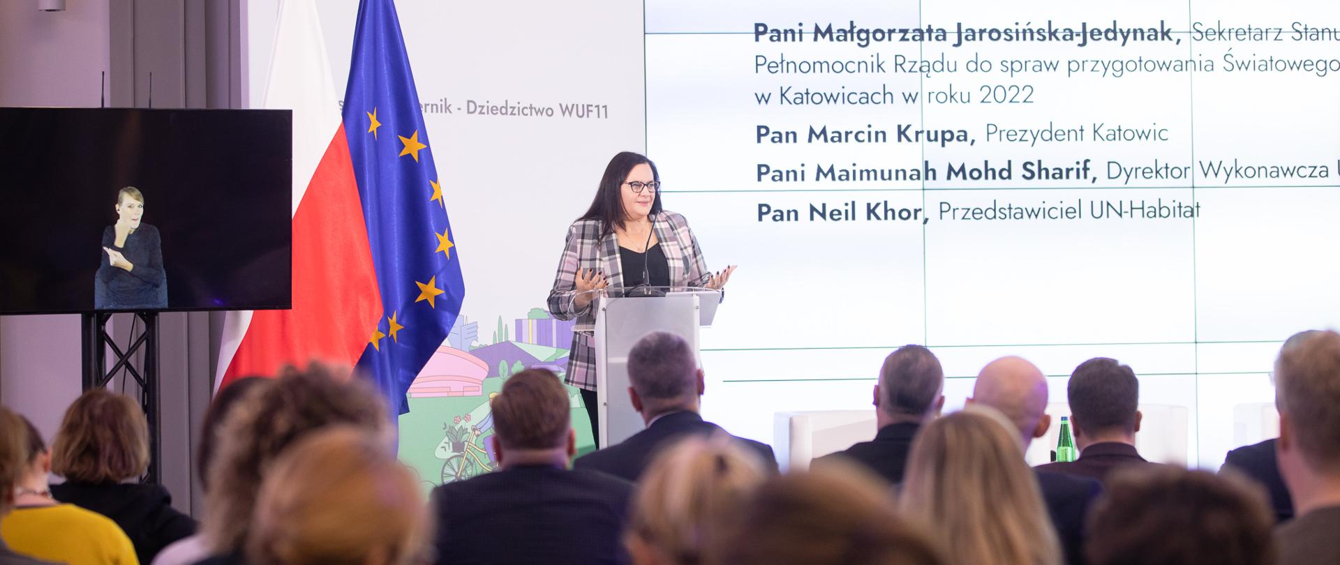 Wiceminister Małgorzata Jarosińska-Jedynak w mównicy na tle flag PL i UE. Przed nią publiczność.