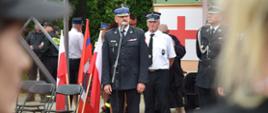 Strażak Państwowej Straży Pożarnej stoi przy mikrofonie. Za nim stoją strażacy ochotnicy w białych koszulach. Przed nim ustawiony jest stojak z trzema flagami oraz krzesła. Dwie skrajne flagi są biało czerwone a środkowa przedstawia znak związku Ochotniczych Straży Pożarnych. W tle na ścianie widać tablicę z umieszczonym na białym tle czerwonym krzyżem.