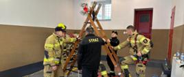 Zdjęcie przedstawia strażaków podczas szkolenia z ratownictwa wysokościowego.
W tle pomieszczenie.
