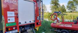 Na zdjęciu widać łódź motorową na przyczepie. Po lewej fragment samochodu ratowniczo-gaśniczego. Samochód czerwony, posiadał białe oznakowanie oraz niebieskie sygnały ostrzegawcze. Zdjęcie wykonane w ciągu dnia. Niebo niebieskie. 