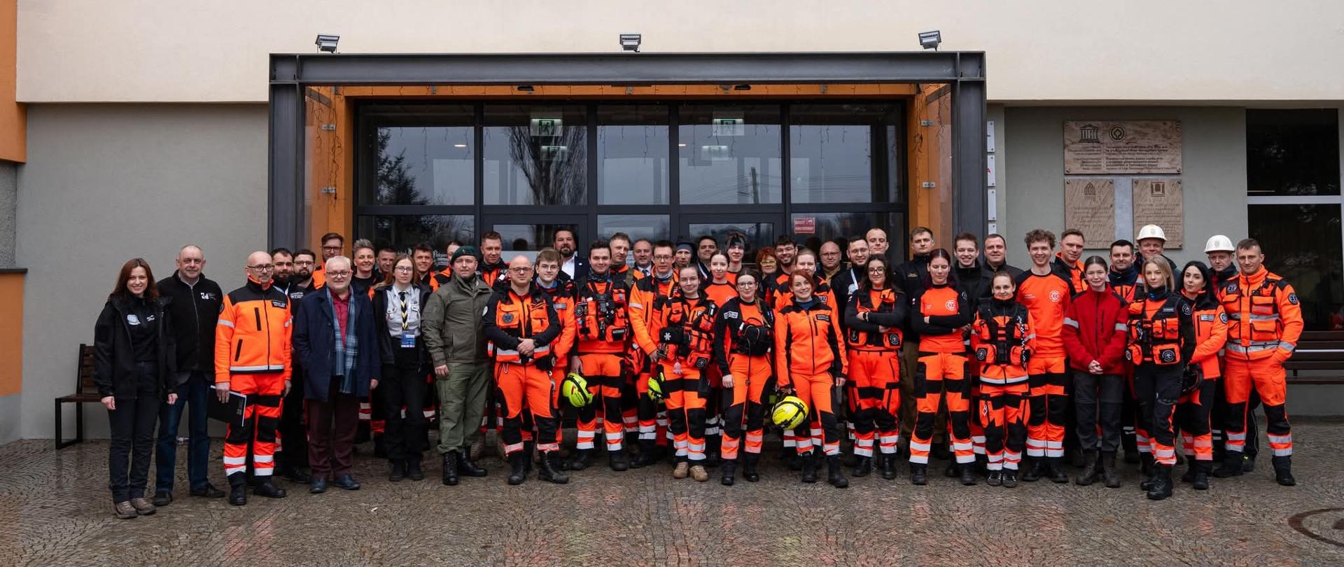 Na zdjęciu znajduje się duża grupa osób pozująca przed wejściem do Zabytkowej Kopalni Srebra w Tarnowskich Górach. Większość zgromadzonych to ratownicy medyczni ubrani w charakterystyczne, jaskrawopomarańczowe uniformy z elementami odblaskowymi i czarnymi wstawkami. Niektórzy z nich trzymają w dłoniach kaski ochronne, a inni mają na sobie odzież cywilną, mundury wojskowe lub stroje harcerskie, co sugeruje wspólne wydarzenie różnych służb i organizacji.
Uczestnicy stoją w kilku rzędach na brukowanym placu przed nowoczesnym, przeszklonym wejściem do budynku. Nad nimi widnieje duży napis z nazwą kopalni, a po prawej stronie drzwi umieszczono pamiątkowe tablice, w tym znak UNESCO. Cała grupa patrzy w stronę obiektywu, tworząc oficjalną, pamiątkową fotografię w pochmurny dzień.